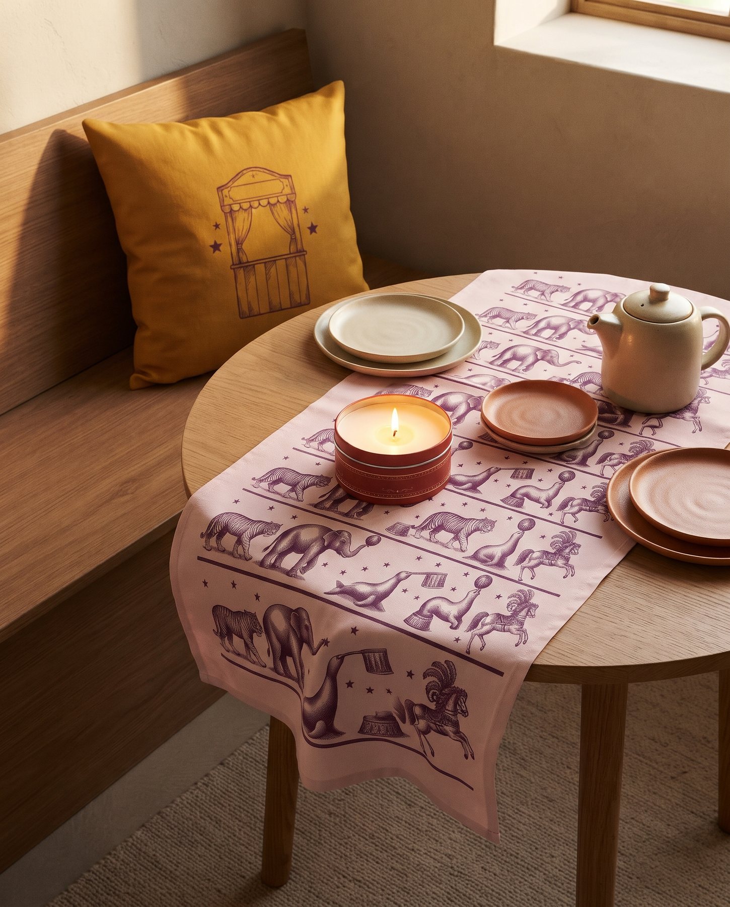A breakfast nook with a marigold circus cushion, a lit red candle tin and a blush menagerie tea towel as a table runner
